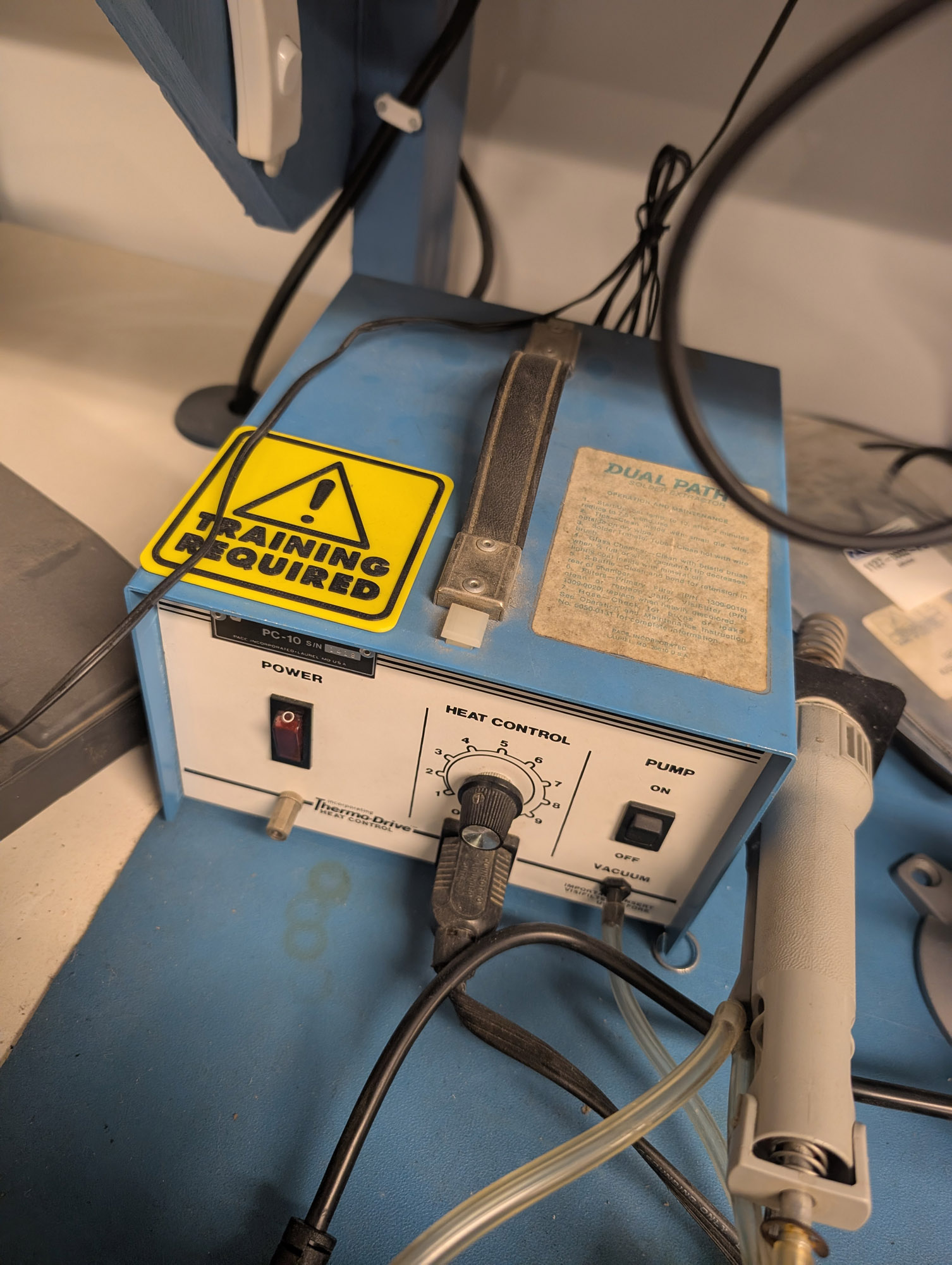 A blue metal box with a white panel on the front. The front has a red illuminated power rocker, a 1-9 Heat Control dial, and a pump on/off switch. Below that is a NEMA 5-15R receptacle, as well as a vacuum connection. The top has a bright yellow "Training Required" sign on it.